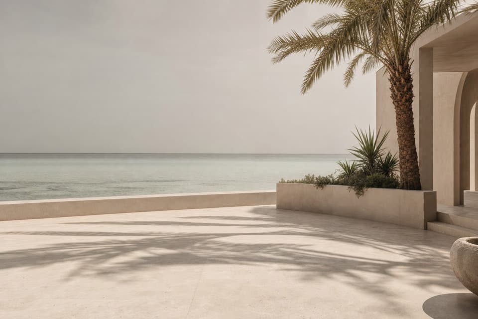 A pale stone terrace in dappled palm shade, opening onto shallow green water under a hazy sky.