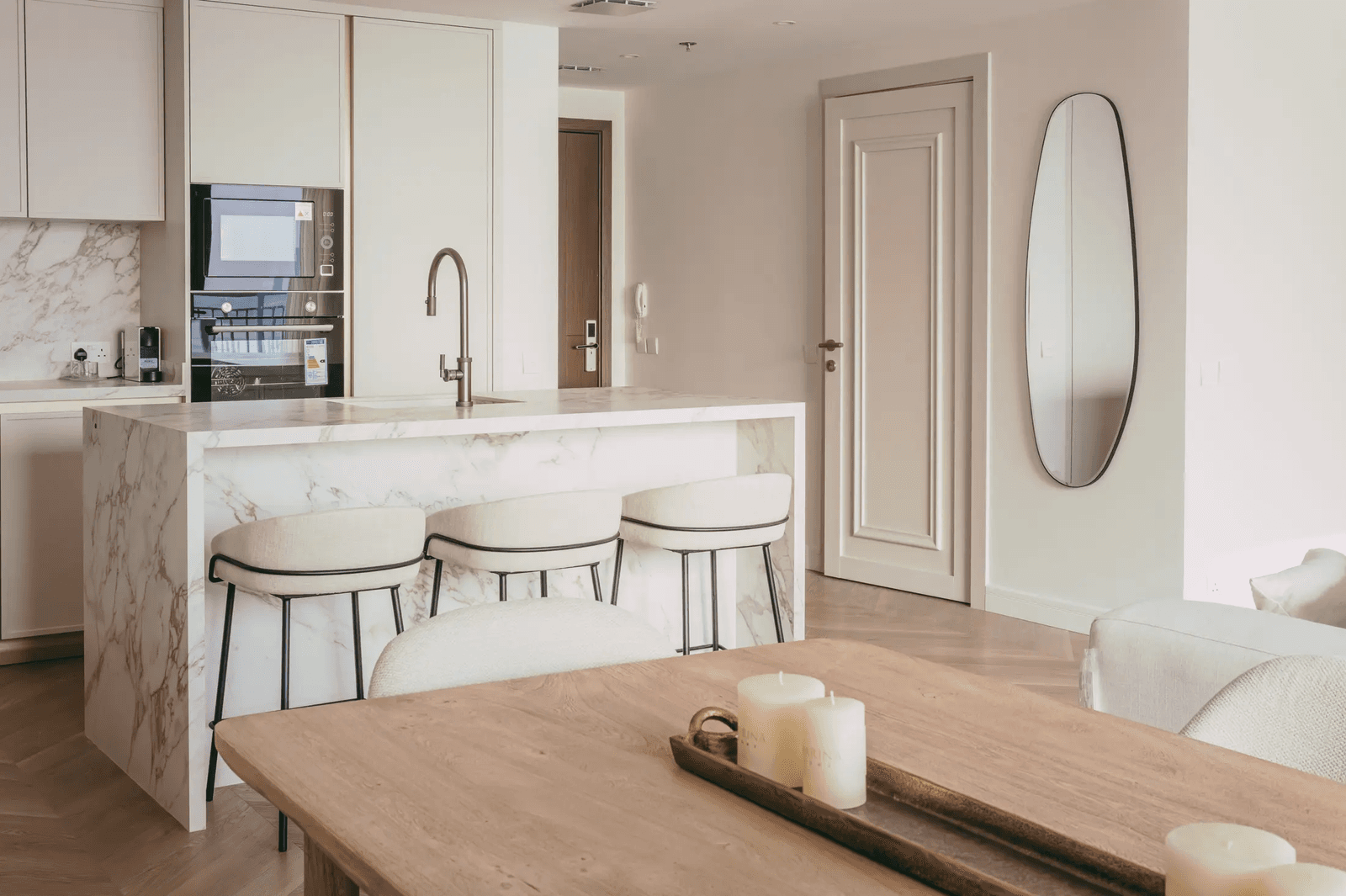 A kitchen island in pale marble with three upholstered stools, seen past a timber dining table.