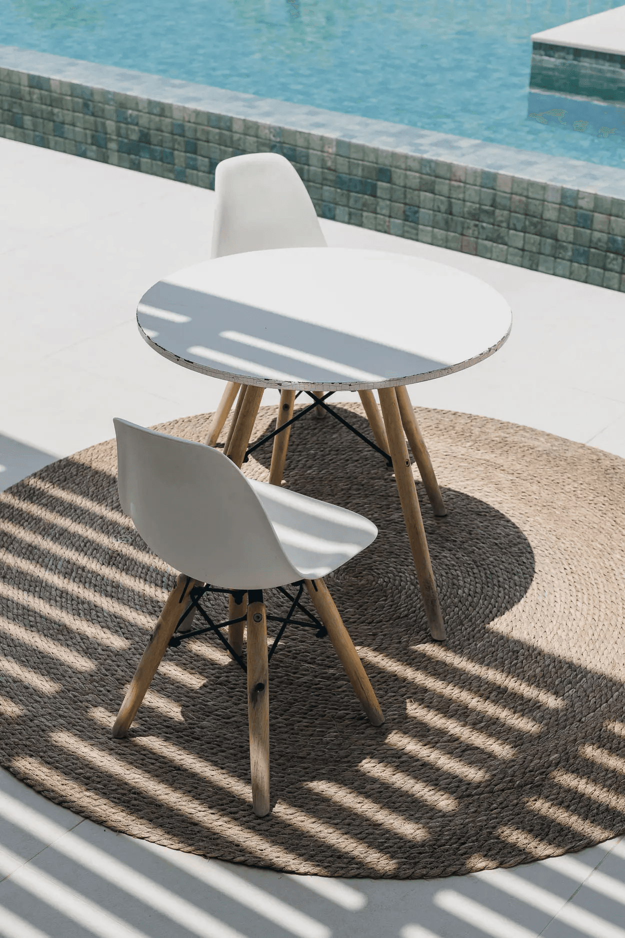 A round white table and two moulded chairs on a woven rug beside a swimming pool.