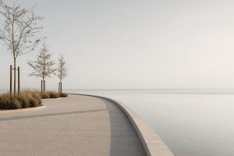 A curving stone promenade edge beside flat still water, with young trees and grasses in morning haze.