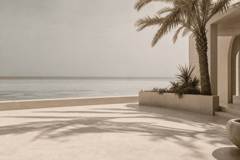 A pale stone terrace in dappled palm shade, opening onto shallow green water under a hazy sky.