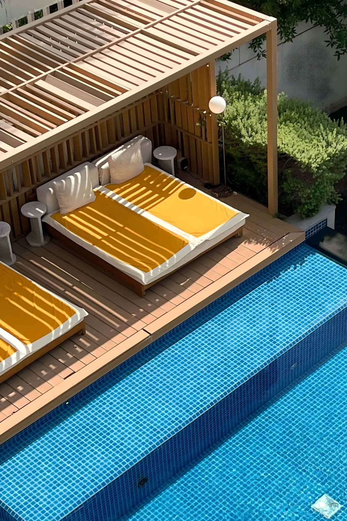 A swimming pool seen from above, beside a timber deck with two daybeds under a slatted pergola.