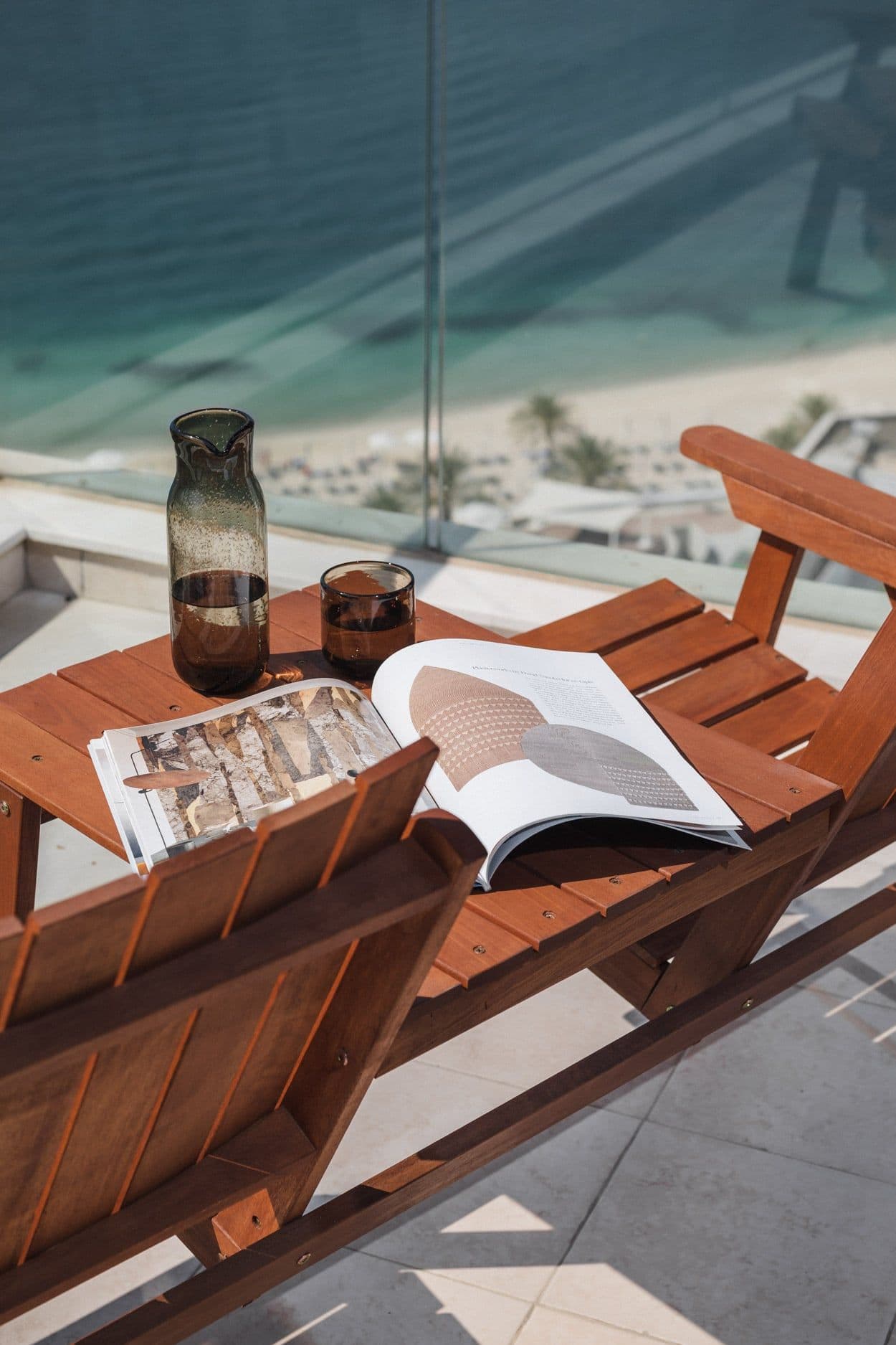 Two timber deck chairs and a table with an open magazine on a balcony high above the beach.