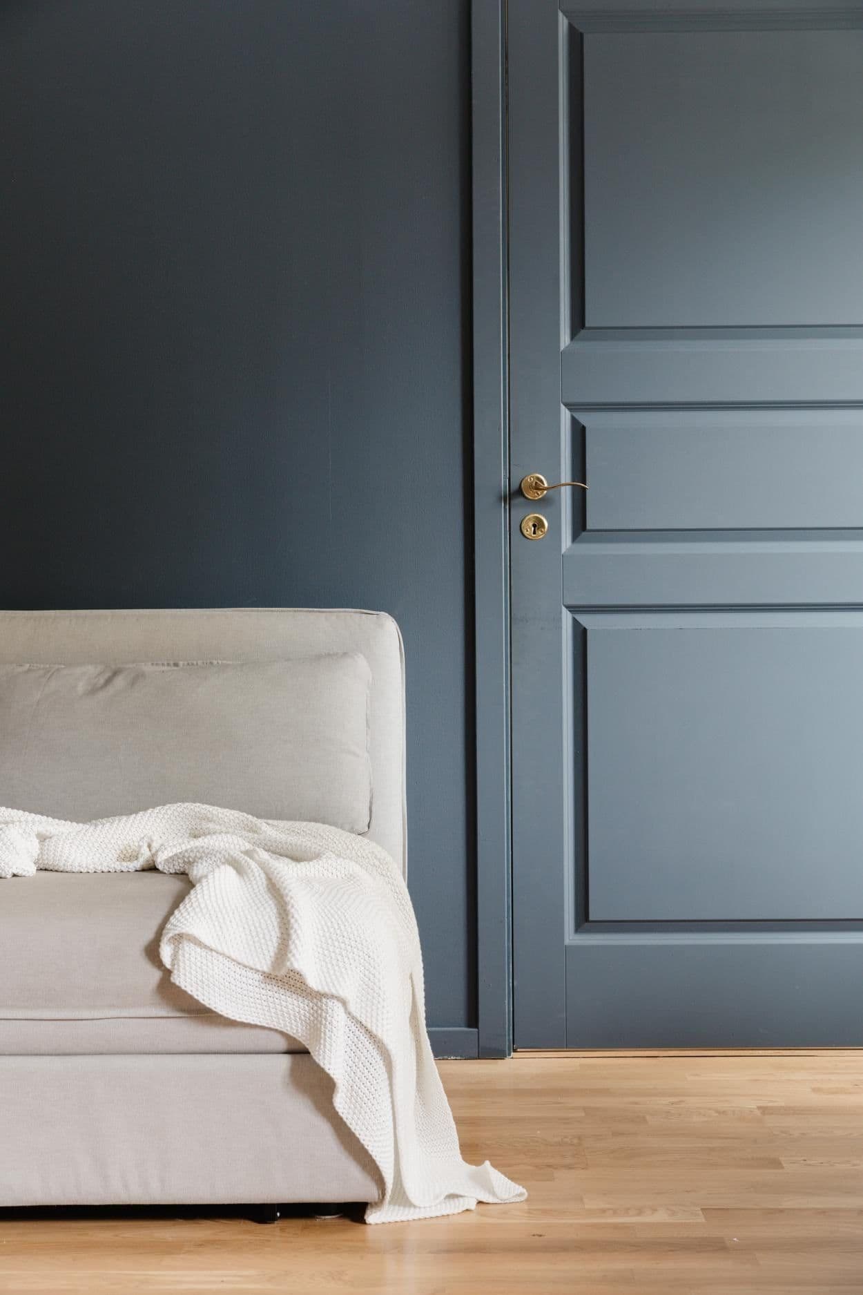 A slate-blue panelled door beside the arm of a pale sofa with a white throw, on an oak floor.