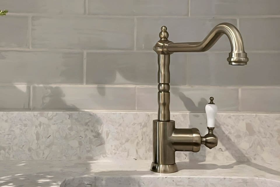 A brushed brass tap on a stone basin, against pale glazed tiles.