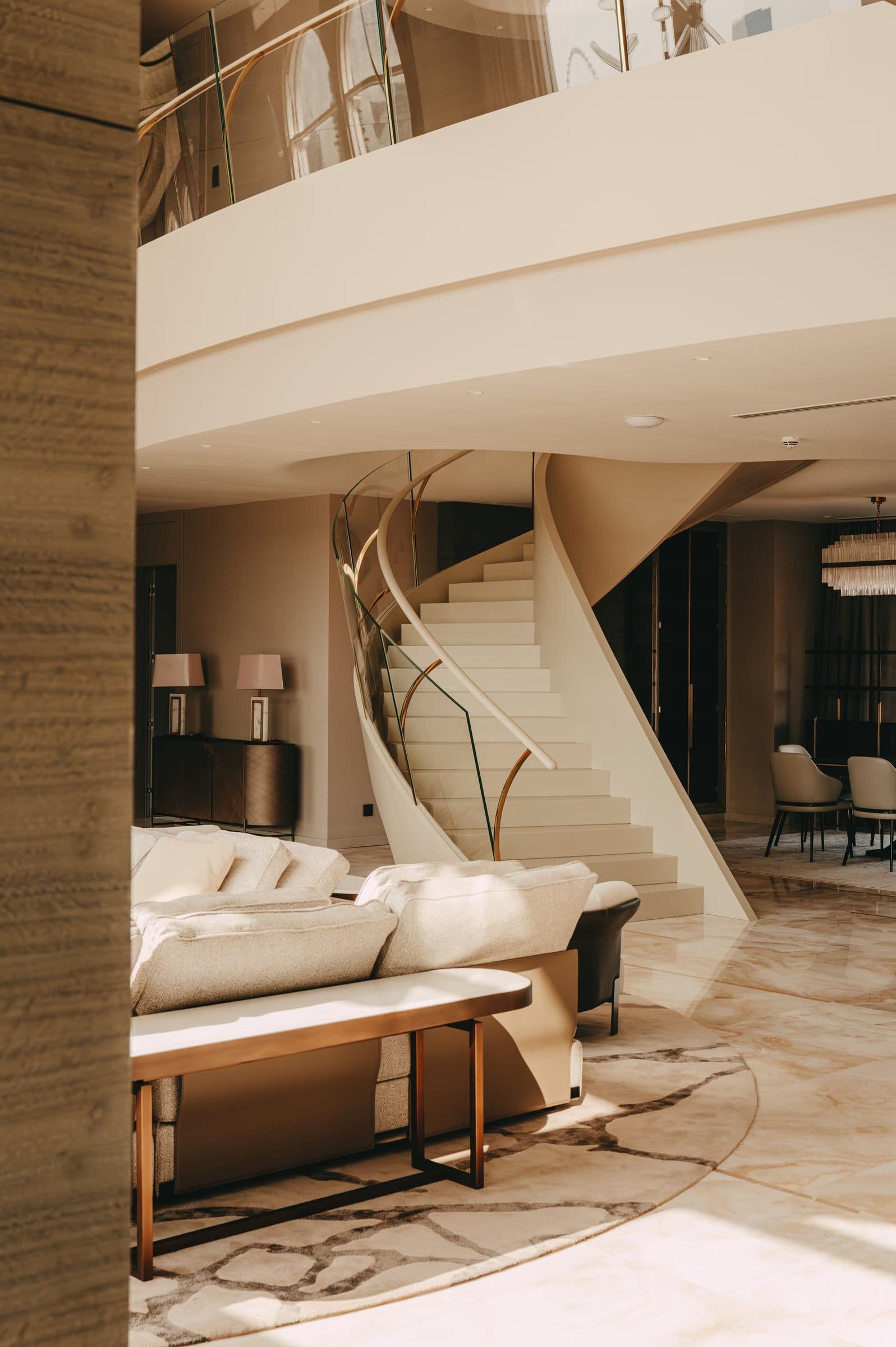 A double-height living room with a curved staircase, a cream sofa and a pale marble floor.