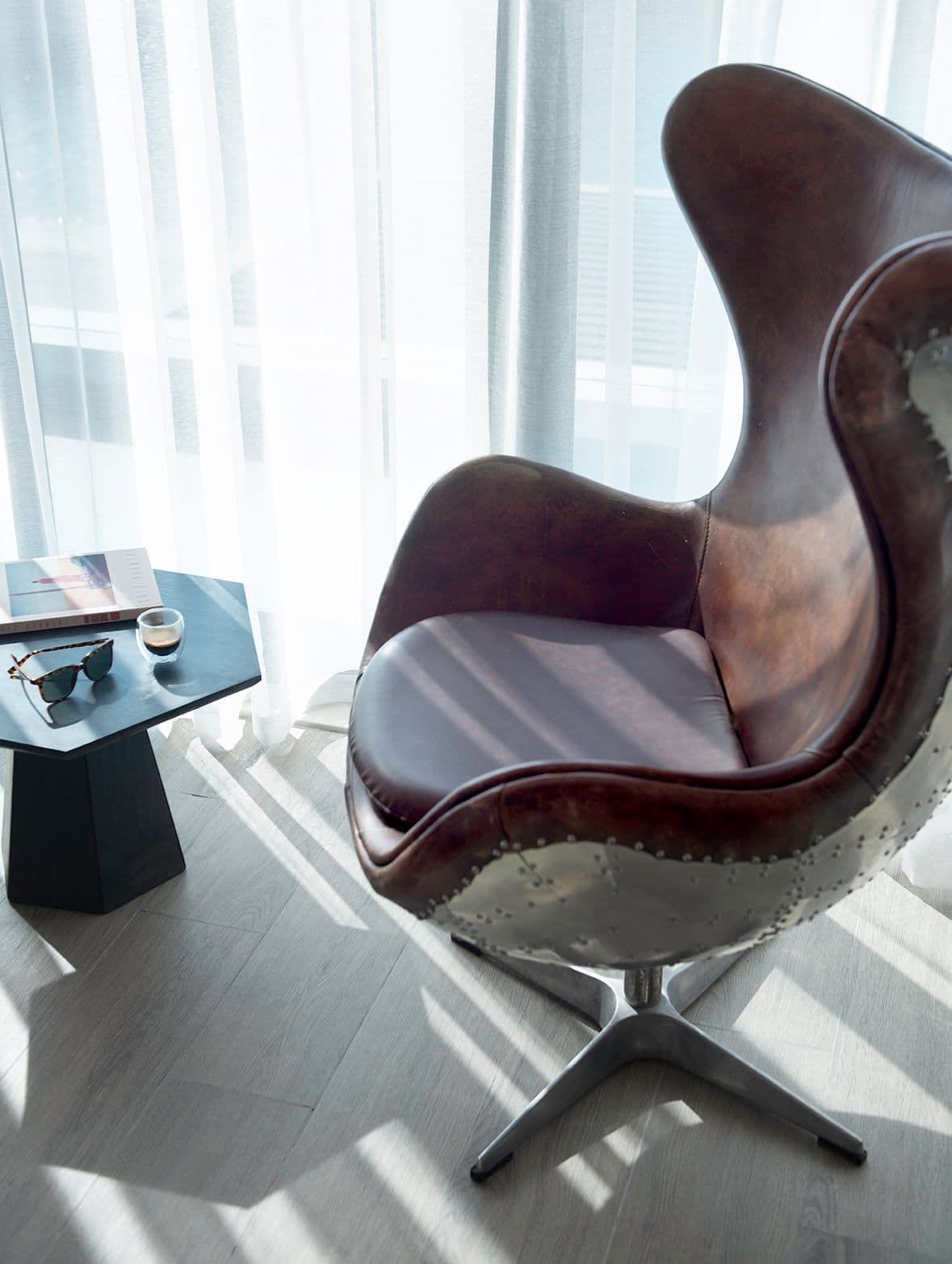 A leather and polished-metal armchair beside a small side table, in sunlight through sheer curtains.