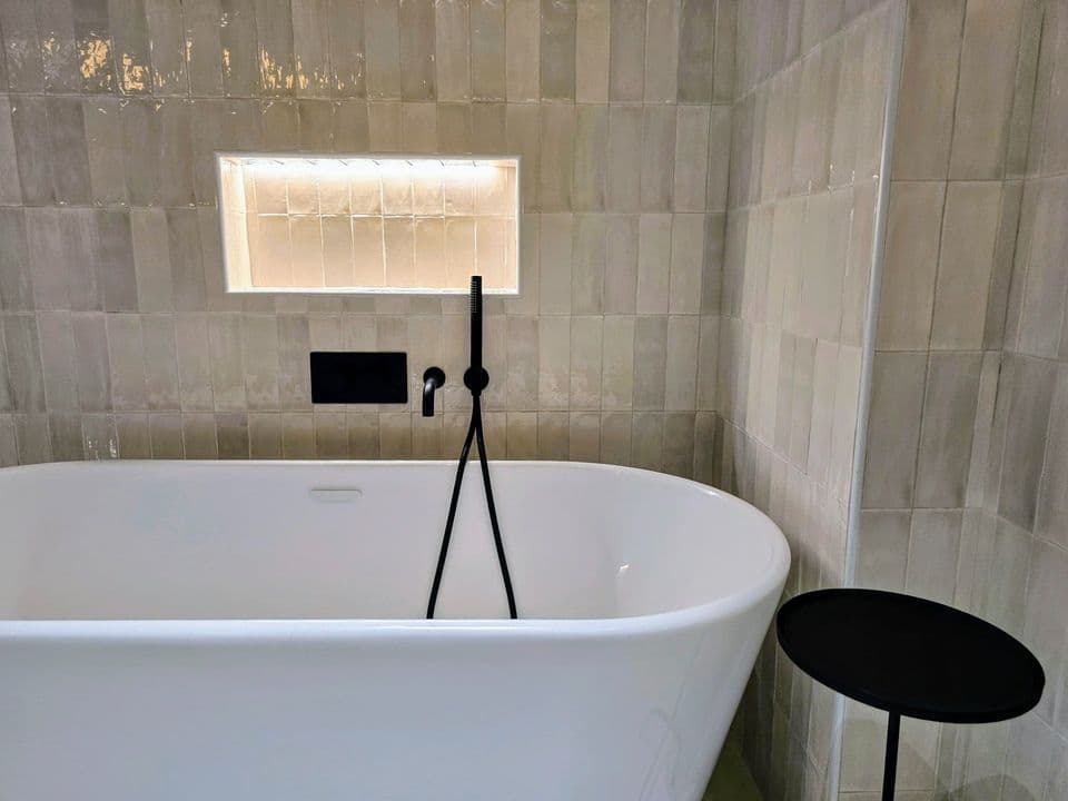 A freestanding bath against handmade grey tiles, with a black floor-standing mixer and a lit niche.