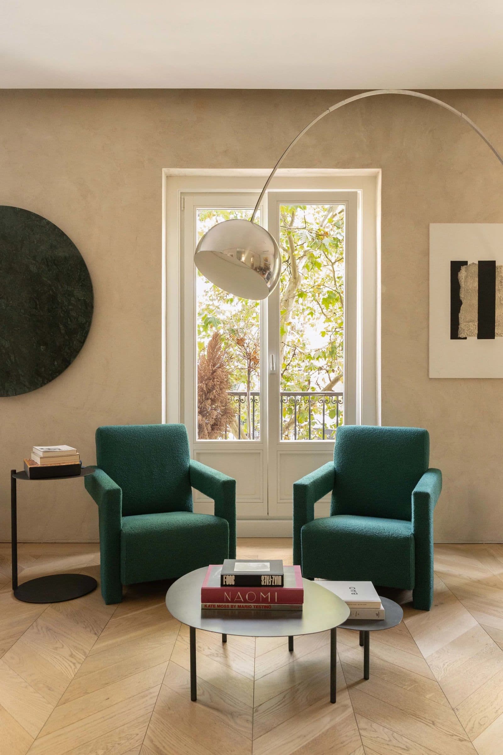 Two green armchairs and a round glass table beneath an arc lamp, in front of a tall window.