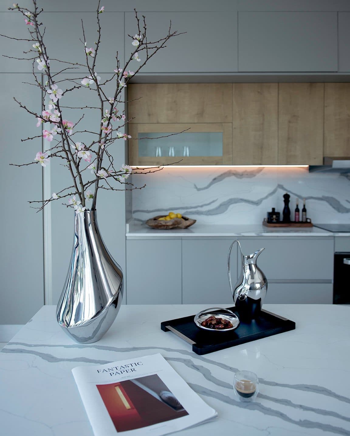 A marble kitchen counter with a silver vase of blossom branches, a tray and a magazine.