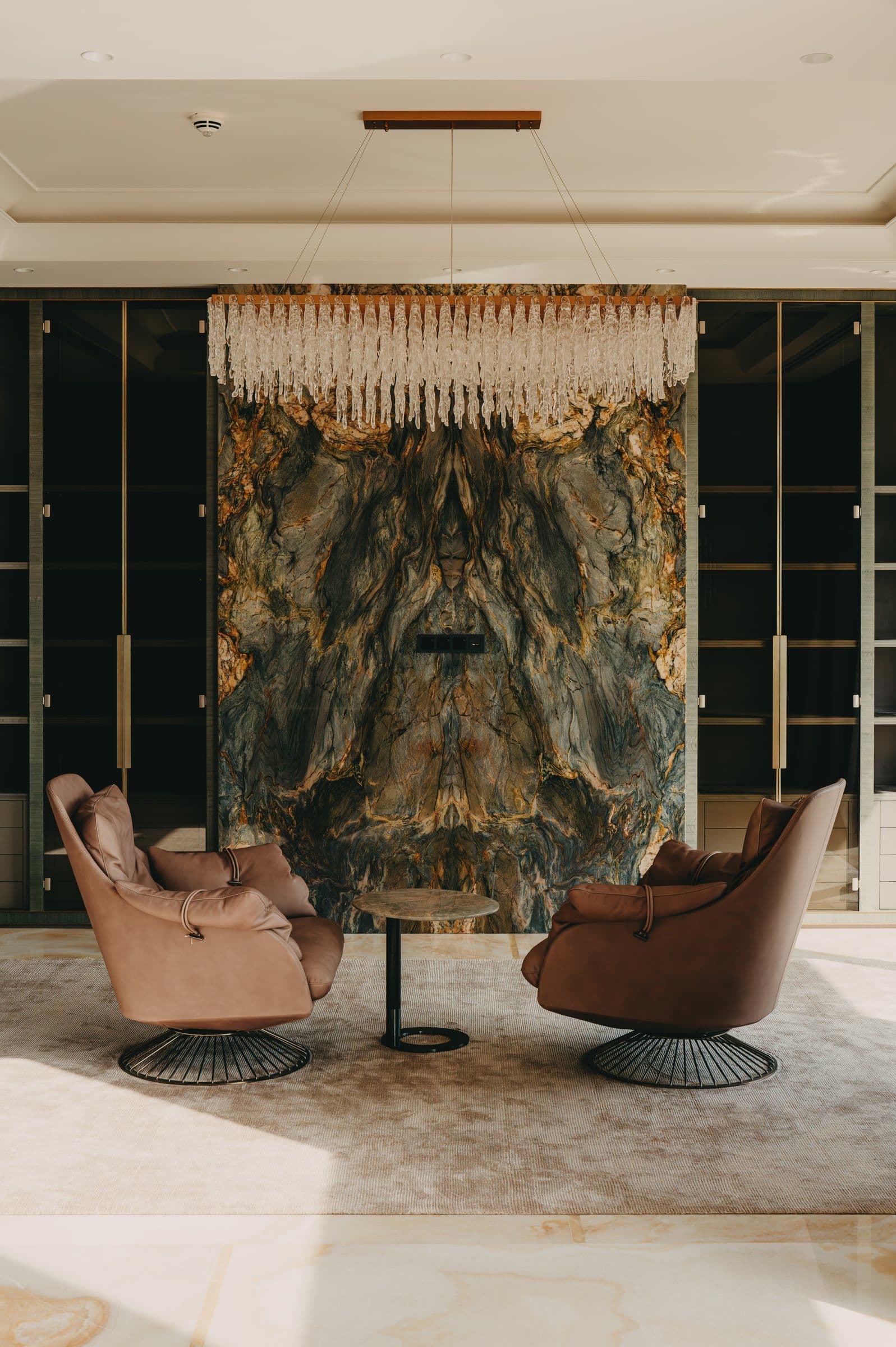 Two leather armchairs beneath a crystal chandelier, in front of a book-matched marble wall.