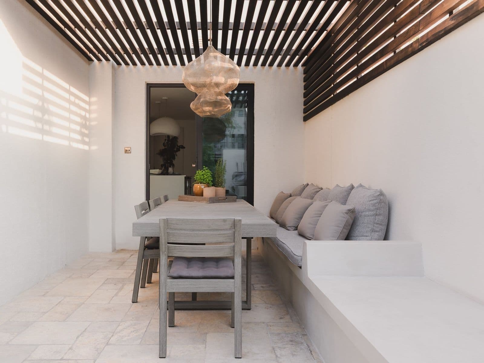 An outdoor dining table under a slatted timber pergola, with a built-in bench and a woven pendant light.
